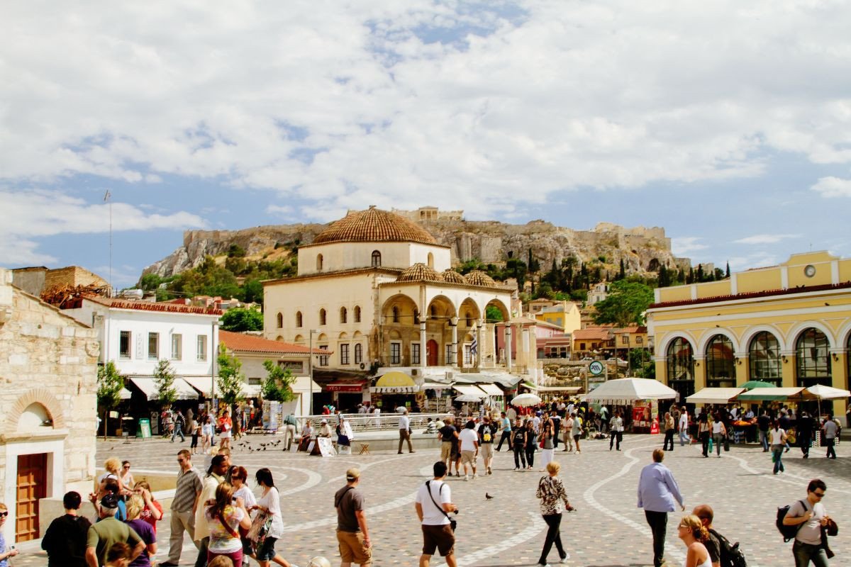 A bustling square in Monastiraki, Athens, filled with people walking and exploring. The scene features historic buildings, including a mosque with a domed roof and arches. In the background, the ancient Acropolis is visible atop a hill, adding a historical and cultural touch to the vibrant urban setting. The sky is partly cloudy, creating a picturesque atmosphere.